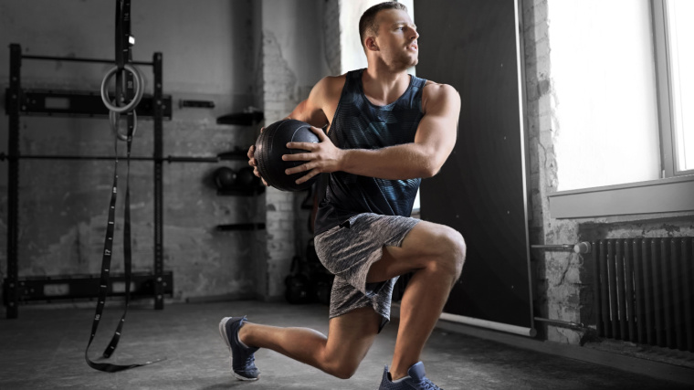 A muscular person doing the lunge exercise while holding a ball in the gym.