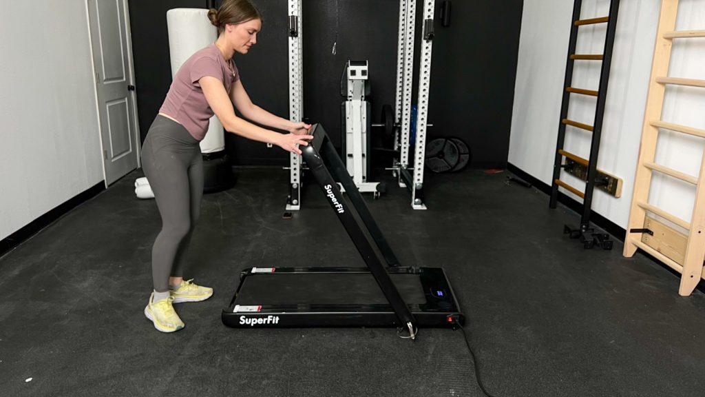 A woman is shown folding the adjustable handlebar on the Goplus 2-in-1 Folding Treadmill.