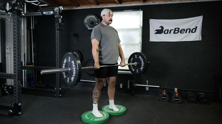 A person wearing a white hat, grey shirt, and black shorts, stands on two green weight plates. They are holding a barbell loaded with a 45-pound pate on either side to perform deficit deadlifts. 