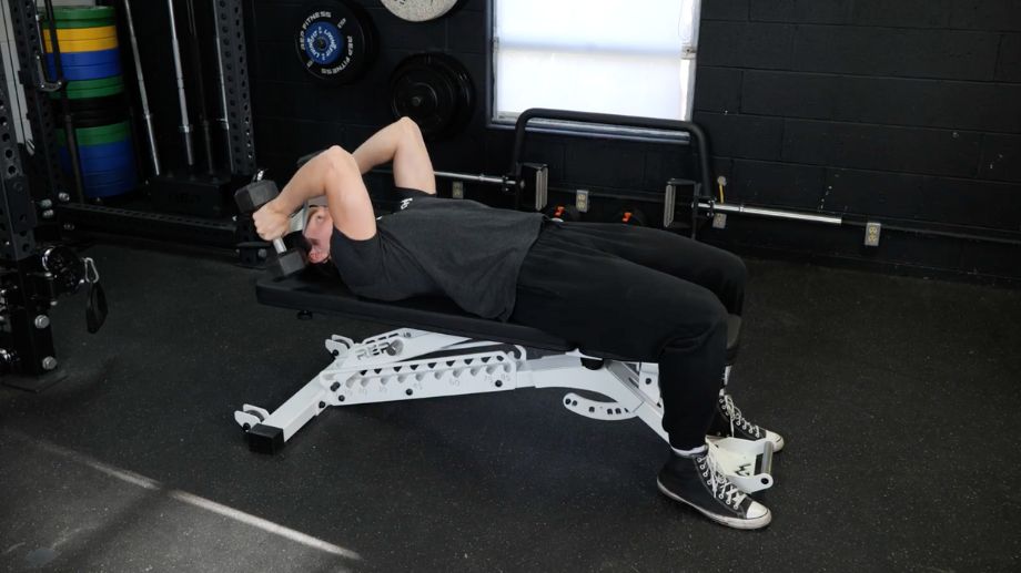 A person wearing black sweatpants and a black t-shirt laying down on a training bench performs dumbbell skull crushers with two hexagonal dummbells.