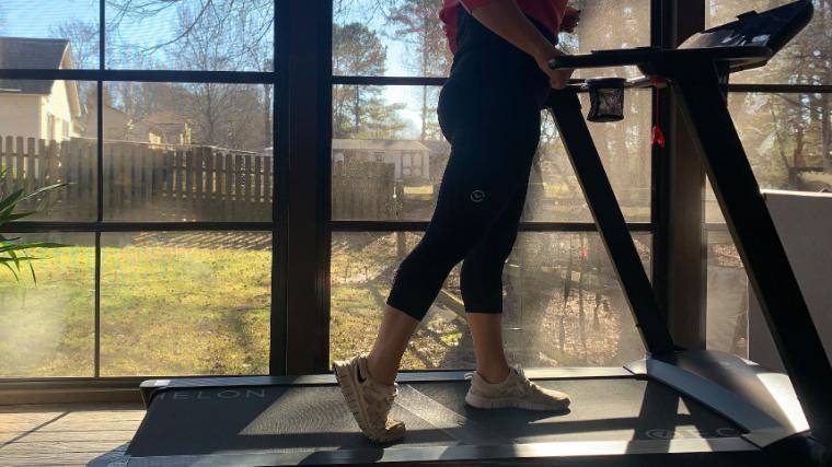 A woman walks on a treadmill with beautiful scenery showing through the windows behind her.