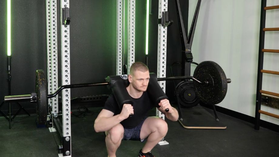 A person wearing blue training shorts and a black t-shirt performs squats with a safety squat bar, loaded with a 25-pound bumper plate on both ends. 