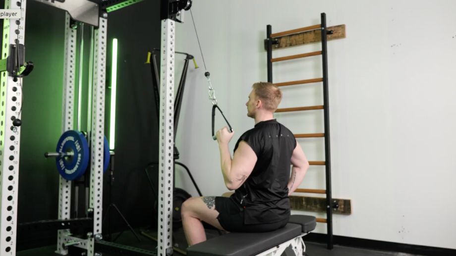A person in a sleeveless workout shirt sits on weight bench - set perpendicular to  a cable pulldown machine. They are performing single-arm cable pulldowns with a D-handle. 
