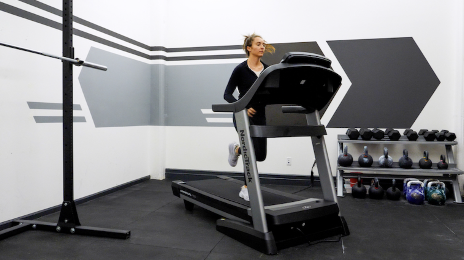 A person in a black long-sleeve shirt runs on a NordicTrack treadmill, set inside a gym. There is a squat rack with a barbell loaded on it in the corner and a rack of dumbbells and kettlebells behind the treadmill.