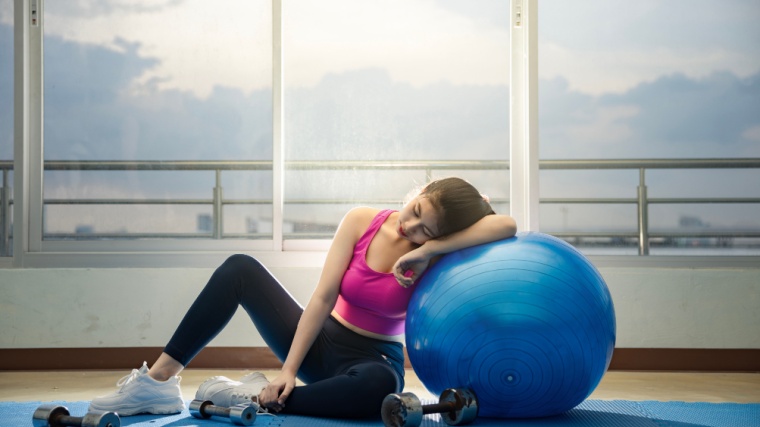 A person who looks tired sitting on an exercise mat, while leaning on an exercise ball.