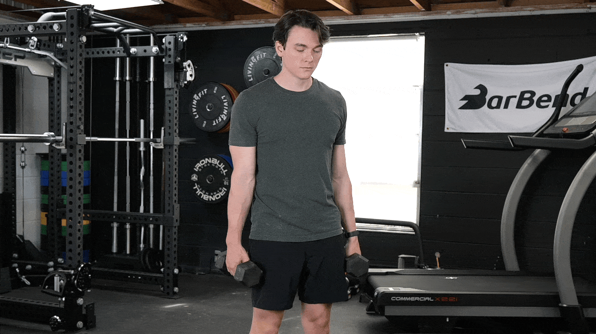 A man wearing a green t shirt with black shorts performs dumbbell wrist curls standing up.