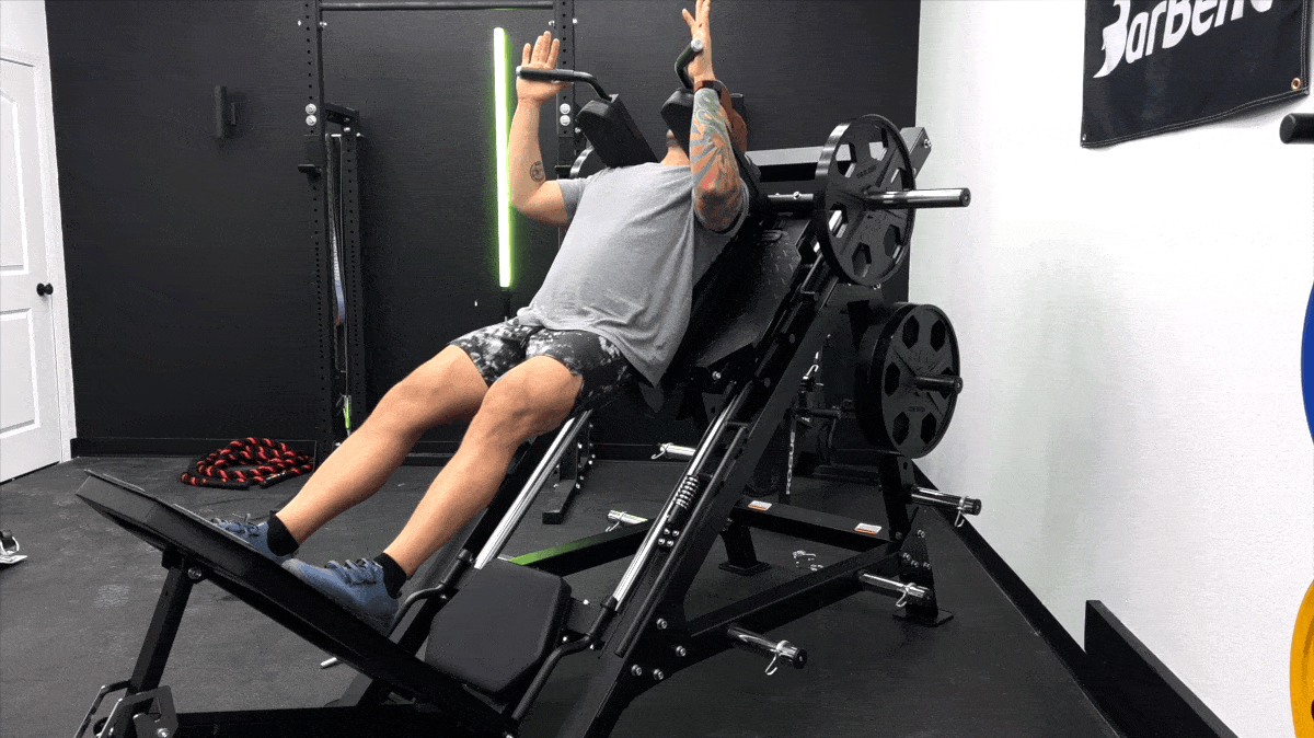 Person wearing a grey shirt and camo pants is positioned vertically in a hack squat machine. They are performing a full-range-of-motion rep, squatting all the way down and exploding back up.