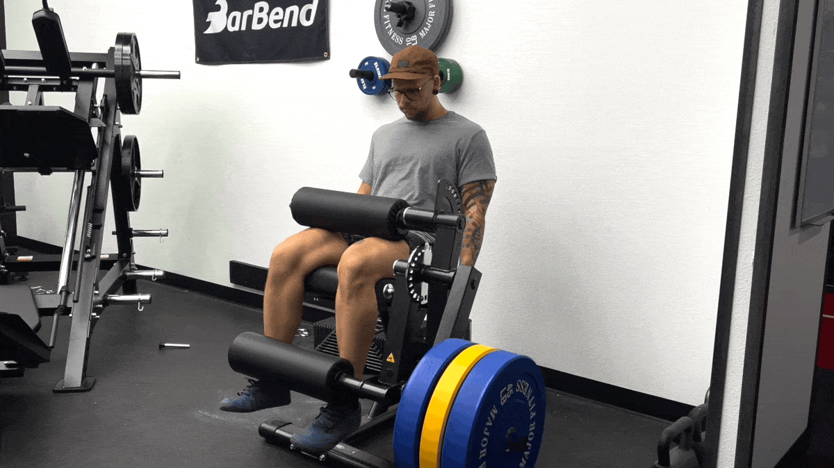 A person wearing a grey t-shirt, shorts, and a brow baseball cap sits in a seated leg extension machine — a pad is secured over their thighs and another pad sits on top of their ankles. They lift their legs to perform a full repetition of the exercise.