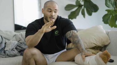 Men's Physique competitor, Jeremy Buendia, sits on a beige sofa with a cast wrapped around his left lower leg. His right hand is raised as he gestures during mid sentence.