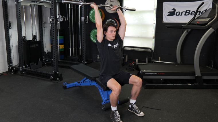 A person holding an EZ bar overhead, while sitting on a weight bench, to do the EZ bar French press exercise.