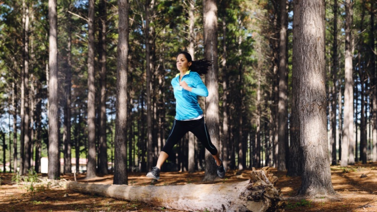 A person trail running in the woods and jumping over a log.