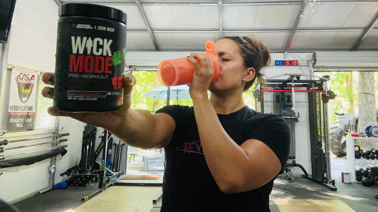 A person drinks from a shaker bottle while holding up a jug of Jacked Factory Wick Mode.
