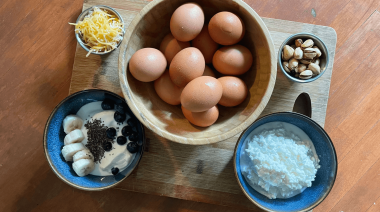 Eggs, yellow cheese, nuts, cottage cheese, and yogurt with some banana, blueberries, and chia seeds sit on a wooden cutting board.