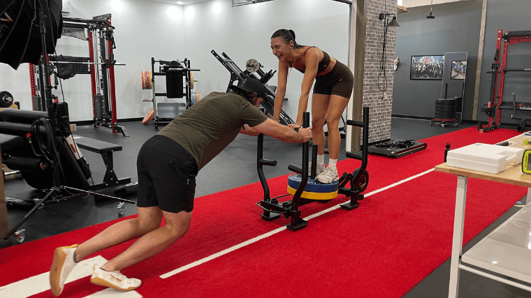 A man pushes the Freak Athlete Wheelbarrow Sled with weight loaded on top, as well as a woman.