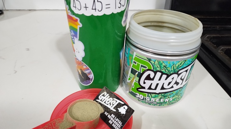 A scoop of Ghost Greens is staged with the opened container and a shaker cup.