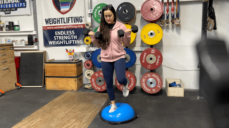 A woman balances on one leg on REP Fitness Balance Ball Trainer while doing bicep curls.